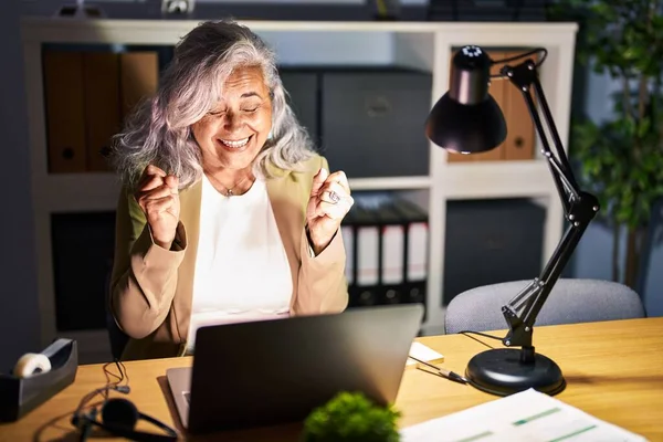 深夜にコンピュータのラップトップを使用して灰色の髪の仕事をしている中年の女性は腕を上げて成功のために興奮し 目を閉じて勝利笑顔を祝う 勝者のコンセプト — ストック写真