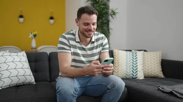 Young Caucasian Man Smiling Using Smartphone Sitting Sofa Home —  Fotos de Stock