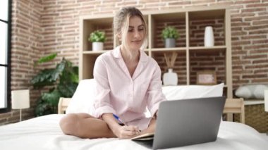Young blonde woman using laptop writing on notebook at bedroom