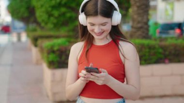 Genç beyaz kadın parkta akıllı telefon ve kulaklık kullanarak gülümsüyor.