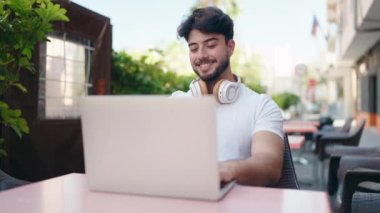 Young hispanic man using laptop sitting on table at coffee shop terrace
