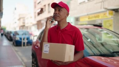 Young latin man delivery worker holding package talking on smartphone at street