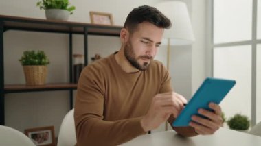 Young hispanic man using touchpad sitting on table at home