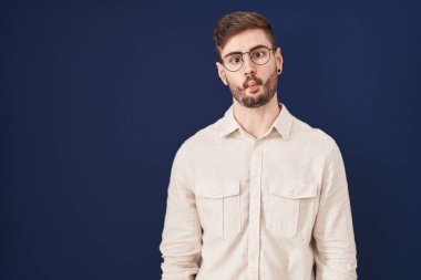 Hispanic man with beard standing over blue background making fish face with lips, crazy and comical gesture. funny expression. 