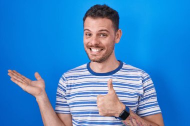 Young hispanic man standing over blue background showing palm hand and doing ok gesture with thumbs up, smiling happy and cheerful 
