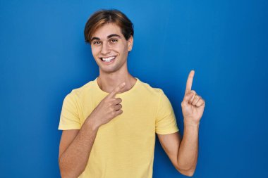 Young man standing over blue background smiling and looking at the camera pointing with two hands and fingers to the side. 