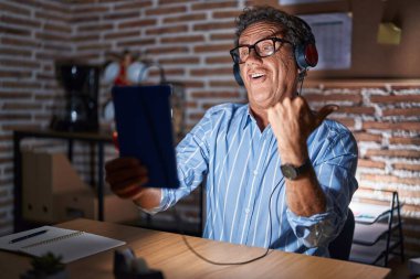 Middle age hispanic man using touchpad sitting on the table at night surprised pointing with hand finger to the side, open mouth amazed expression. 