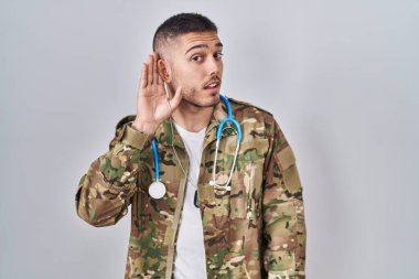Young hispanic doctor wearing camouflage army uniform smiling with hand over ear listening and hearing to rumor or gossip. deafness concept. 
