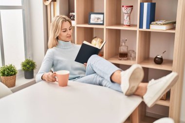 Genç sarışın kadın kitap okuyor ve evde kahve içiyor.