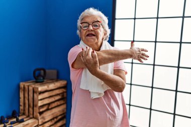 Spor kıyafetleri giyen yaşlı gri saçlı kadın spor merkezinde kendine gülerek duruyor.