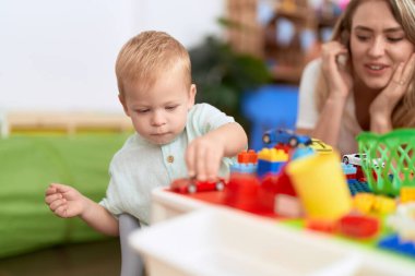 Anaokulunda oturan öğretmen ve yeni yürümeye başlayan çocuk inşaat blokları ve araba oyuncaklarıyla oynuyor.