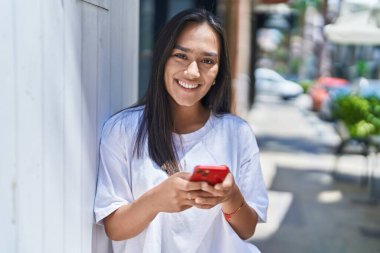 Genç, güzel, İspanyol bir kadın sokakta akıllı telefon kullanarak gülümsüyor.