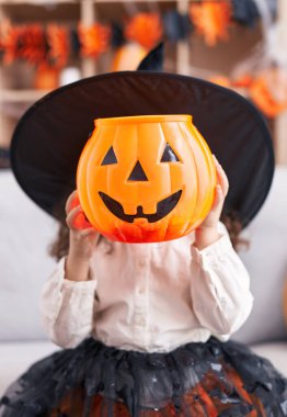 Adorable hispanic girl wearing halloween costume holding pumpkin basket over face at home