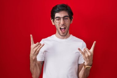 Young hispanic man standing over red background shouting with crazy expression doing rock symbol with hands up. music star. heavy music concept. 