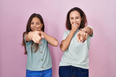 Young mother and daughter standing over pink background laughing at you, pointing finger to the camera with hand over mouth, shame expression 