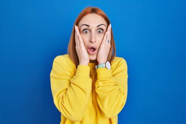 Young woman standing over blue background afraid and shocked, surprise and amazed expression with hands on face 