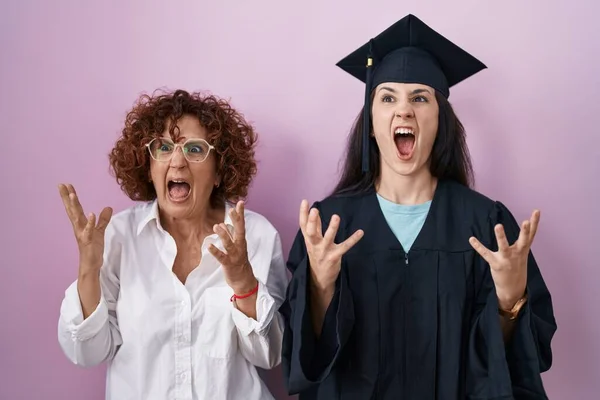 卒業キャップと式のローブを身に着けているヒスパニック系の母親と娘は狂った叫びと積極的な表現と腕を上げて叫んでいます フラストレーションコンセプト — ストック写真
