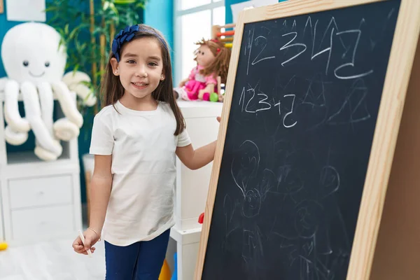 在幼儿园的黑板上 他那可敬的惊慌失措的学龄前女生微笑着写下了自信的数字 — 图库照片