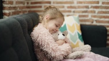 Adorable blonde girl hugging teddy bear sitting on sofa at home
