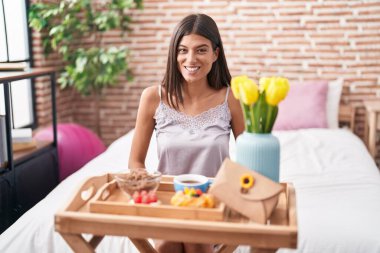 Brunette young woman eating breakfast sitting on the bed looking positive and happy standing and smiling with a confident smile showing teeth 