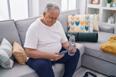 Orta yaşlı, kır saçlı adam evde elinde bir şişe su tutan akıllı telefon kullanıyor.