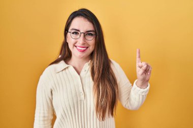 Sarı arka planda duran genç İspanyol kadın bir numarayı gösterip işaret ederken kendinden emin ve mutlu bir şekilde gülümsüyor.. 