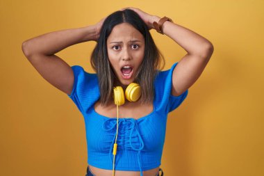 Hispanic young woman standing over yellow background crazy and scared with hands on head, afraid and surprised of shock with open mouth 