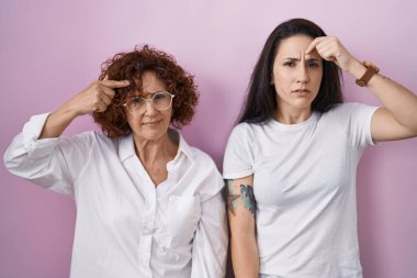 İspanyol anne ve kızı pembe arka plan üzerine gündelik beyaz tişört giyiyorlar. Alnındaki sivilceden mutsuz olduklarını gösteriyorlar. Siyah noktalı çirkin bir enfeksiyon. sivilce ve deri problemi 