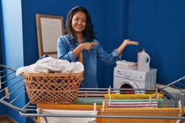 Young asian woman hanging clothes at clothesline amazed and smiling to the camera while presenting with hand and pointing with finger. 