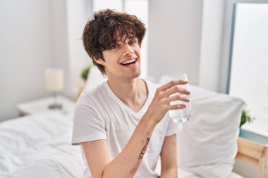 Young hispanic man drinking glass of water sitting on bed at bedroom