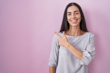 Genç esmer kadın pembe arka planda neşeli bir şekilde ayakta duruyor. Yüzünde mutlu ve doğal bir ifadeyle elleri ve parmaklarıyla gülümsüyor. 