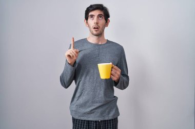 Genç İspanyol adam pijama giyip bir fincan kahve içerken şaşkın şaşkın yukarı bakıyor ve parmaklarıyla işaret ediyor ve kollarını kaldırıyor.. 