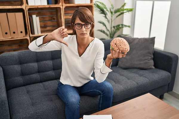 Brunette woman working at therapy office holding brain with angry face, negative sign showing dislike with thumbs down, rejection concept