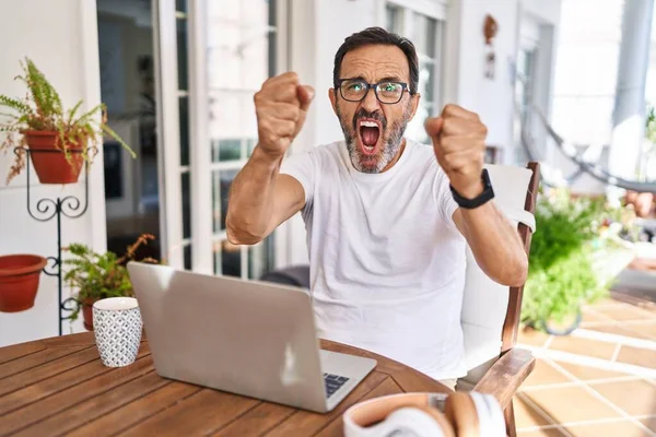 Middle age man using computer laptop at home angry and mad raising fists frustrated and furious while shouting with anger. rage and aggressive concept.