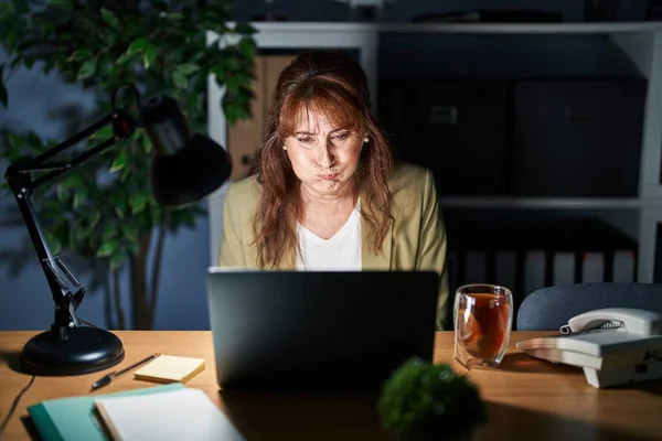 Middle age hispanic woman working using computer laptop at night puffing cheeks with funny face. mouth inflated with air, crazy expression.