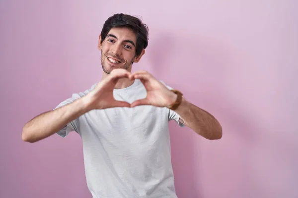 Jonge Spaanse Man Staat Roze Achtergrond Glimlachend Liefde Doen Hart — Stockfoto