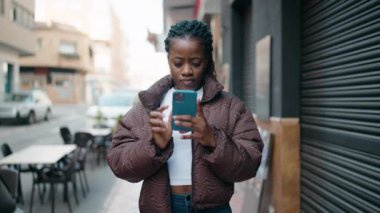 Afro-Amerikalı kadın gülümsüyor. Sokakta akıllı telefonun yanında özçekim yapıyor.