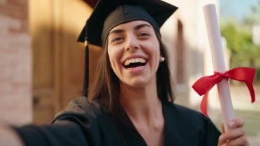 Mezuniyet üniforması giyen genç İspanyol kadın elinde diplomayla üniversitede video görüşmesi yapıyor.