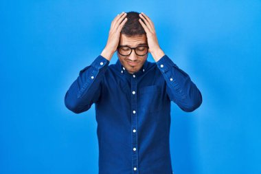 Young hispanic man wearing glasses over blue background suffering from headache desperate and stressed because pain and migraine. hands on head. 