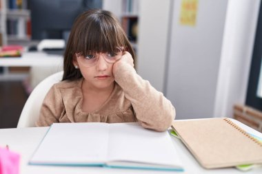 Adorable hispanic girl student writing on notebook with relaxed expression at classroom