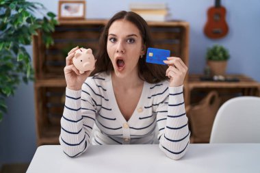 Young hispanic girl holding piggy bank and credit card afraid and shocked with surprise and amazed expression, fear and excited face. 