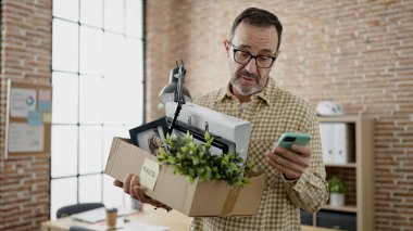 Middle age man business worker dismissed holding cardboard box using smartphone at office