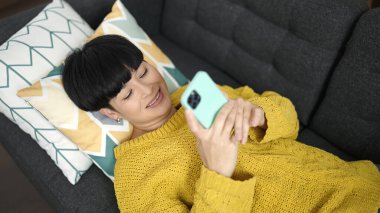 Young chinese woman using smartphone lying on sofa at home