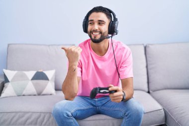 Hispanic young man playing video game holding controller sitting on the sofa smiling with happy face looking and pointing to the side with thumb up. 