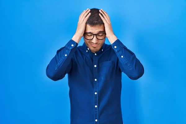 痛みや片頭痛のために絶望的な頭痛やストレスに苦しんで青い背景に眼鏡をかけて若いヒスパニック系の男 手を頭に — ストック写真