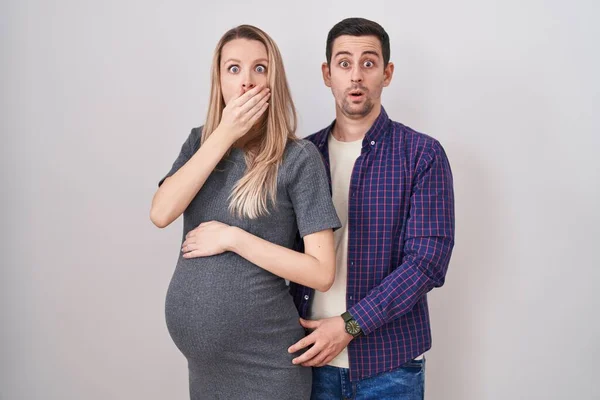 白い背景の上に立つ赤ん坊を期待している若いカップルは間違いのための手で口を覆うショックを受けた 秘密の概念 — ストック写真