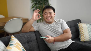 Young chinese man using smartphone doing ok gesture at home