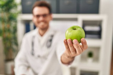 Diyetisyen üniforması giyen genç İspanyol adam klinikte elma tutuyor.