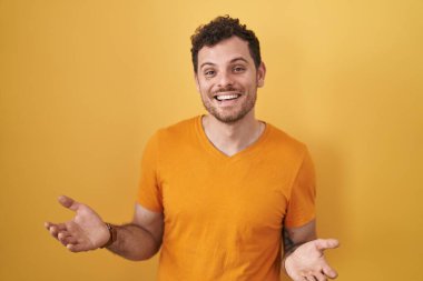 Young hispanic man standing over yellow background smiling cheerful with open arms as friendly welcome, positive and confident greetings 