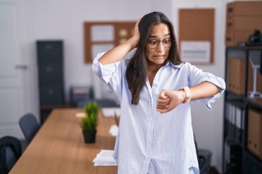 Young hispanic woman at the office looking at the watch time worried, afraid of getting late 